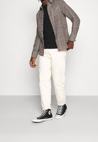 Hombre con pantalones color crema, zapatillas negras de caña alta, camiseta negra y camisa de cuadros desabrochada, de pie contra un fondo simple.