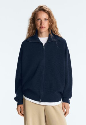 Vrouw met golvend blond haar, gekleed in een marineblauwe trui met ritssluiting over een wit shirt en beige broek, staand tegen een effen achtergrond.