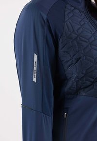 Giacca blu navy con texture trapuntata sulla parte superiore del petto, corpo liscio e logo riflettente "Swedemount". Presenta una tasca laterale con zip.