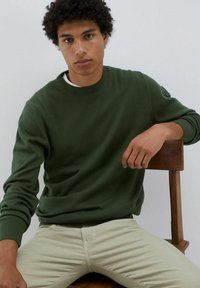 Maglione verde lavorato a maglia con scollatura tonda e polsini a costine, abbinato a pantaloni beige chiaro, seduto su una sedia di legno contro uno sfondo semplice.