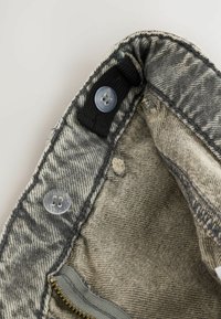 Parte interna della vita di un paio di jeans di jeans grigi che mostrano una fascia elastica nera regolabile con bottoni trasparenti e una cerniera parzialmente visibile.