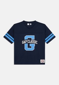 Tumši zils sieta īsās piedurknes T-krekls ar V-veida kakla izgriezumu, ar gaiši ziliem svītrām uz piedurknēm un "GAP CLASSIC" grafiku baltā un zilā krāsā.