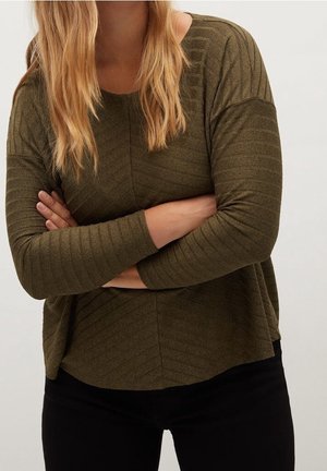 Osoba z długimi blond włosami, ubrana w oliwkowozielony prążkowany top z długim rękawem i czarne spodnie, z założonymi rękami na piersi.