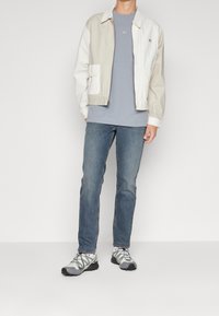 Lättviktsjacka i färgblock, beige och vit, tillsammans med en grå t-shirt, blå jeans och grå sportskor med svarta detaljer.