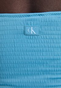 Turkizna tkanina z žlebasto strukturo, ki vključuje majhno modro oznako z belo "cK" logotipom. Gladek, raztegljiv material.