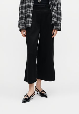 Osoba nosí čierne široké krátke nohavice, čierne špicaté slingbackové lodičky s mašľami, čierne tričko a čierno-biely kockovaný kabát.