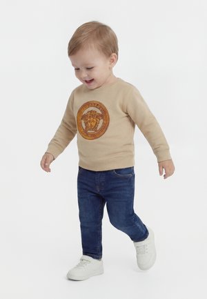 Mažas vaikas su šviesiai rudais plaukais vaikšto, dėvi smėlio spalvos džemperį su apskritu emblematu, tamsiai mėlynas džinsus ir baltus sportinius batelius ant baltos spalvos fono.