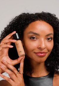 Femme aux cheveux bouclés tient un flacon de fond de teint KIKO Milano près de son visage, souriant doucement devant un fond neutre.