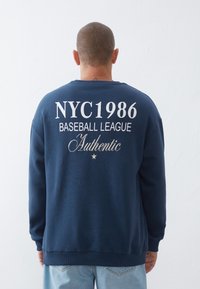 Felpa blu navy con maniche lunghe, collo rotondo e testo bianco sulla parte posteriore: "NYC 1986 BASEBALL LEAGUE Authentic" con un dettaglio a stella.