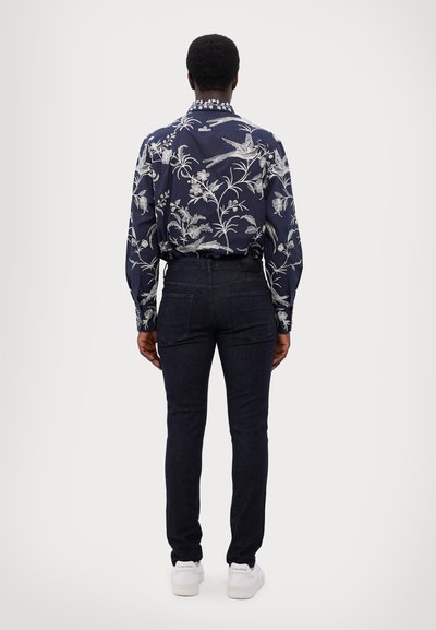 Camisa de patrón floral en tonos navy con acentos blancos, con cuello abotonado, combinada con jeans oscuros y zapatillas blancas, vista desde la parte de atrás.