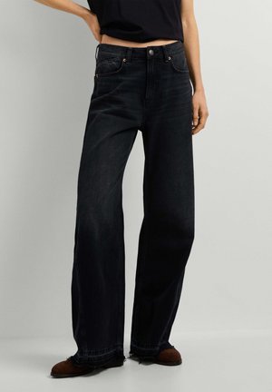Personne portant un jean noir taille haute à jambes larges avec des ourlets effilochés et des chaussures marron, debout devant un fond clair uni.