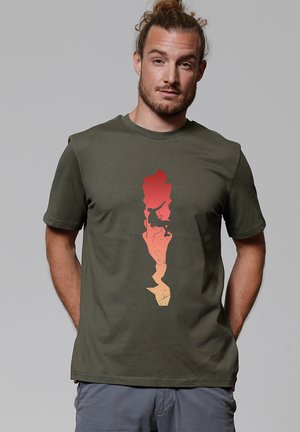 Mann mit zurückgebundenen Haaren, der ein olivgrünes T-Shirt mit einem rot-gelb verlaufenden Kletterer-Motiv trägt, die Hände in den Taschen.