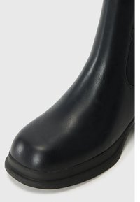 Botín negro de tobillo con material de cuero sintético suave, punta redonda y una suela de goma delgada; cuenta con paneles elásticos para facilitar su uso.