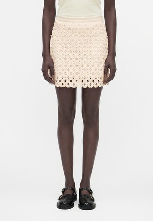 Mini sukňa v krémovej farbe s geometrickým vzorom výrezov, nosená s čiernymi Mary Jane topánkami na pracku a bezrukávovým topom.