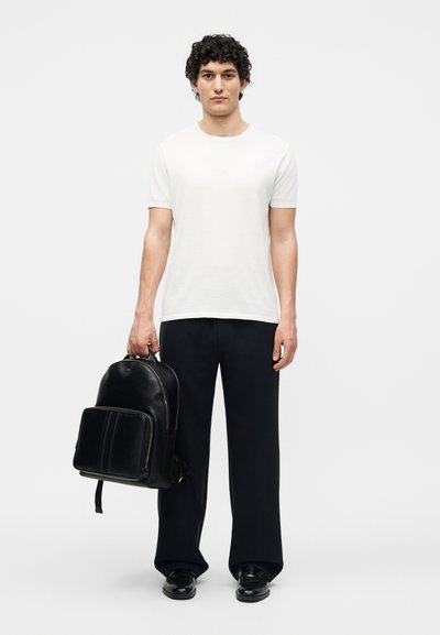 Homme aux cheveux bouclés portant une chemise blanche à manches courtes, un pantalon large noir, des chaussures noires, tenant un sac à dos en cuir noir à ses côtés.