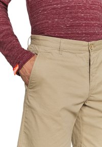 Pantalon beige en tissu texturé, avec une fermeture à bouton, des poches latérales et une étiquette de marque rouge visible sur le côté gauche.