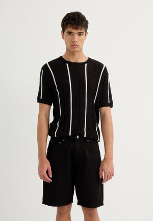 Camisa negra de punto de manga corta con rayas verticales blancas, combinada con pantalones cortos de mezclilla negros. El modelo está de pie contra un fondo sencillo.