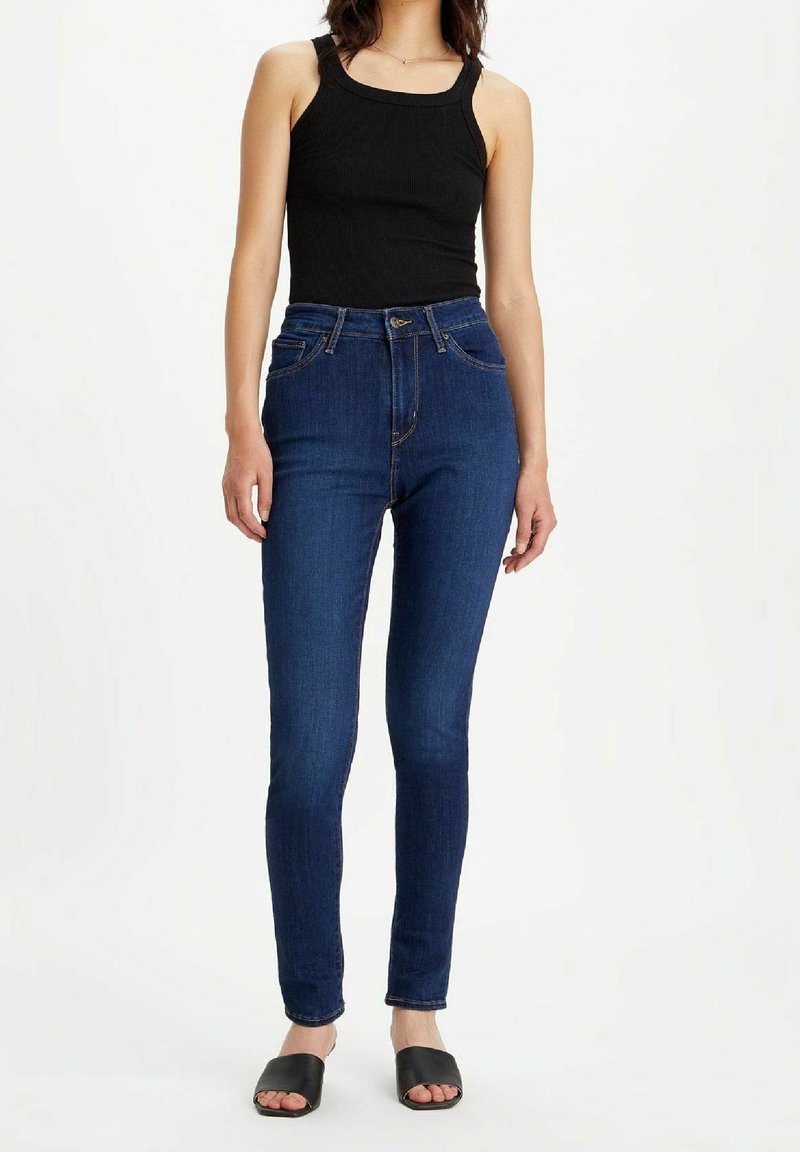 Femme portant un haut noir sans manches, un jean skinny bleu foncé taille haute et des chaussures noires ouvertes à enfiler, debout devant un fond blanc.