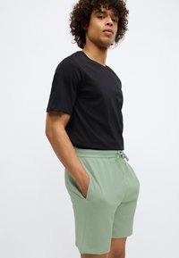 Tricou negru cu mânecă scurtă, asortat cu pantaloni scurți texturați de culoare verde deschis, care au buzunare laterale și un talie cu șnur.