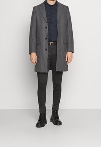 Manteau gris en mélange de laine avec revers crantés, coupe à simple boutonnage, associé à un pantalon sombre ajusté et des bottines noires à lacets.