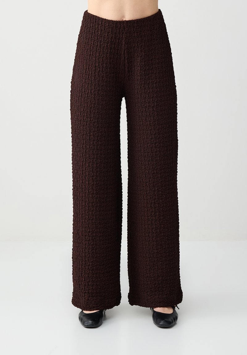 Pantalon large texturé marron foncé porté avec des chaussures plates noires par une personne debout devant un fond uni.
