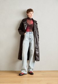 Cappotto di pelle marrone lungo su un top trasparente rosso e nero, abbinato a jeans azzurri a vita alta e scarpe marroni e rosse.