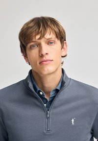 Grijze rits-trui met kraag, voorzien van een wit logo op de linkerkant van de borst. Onderneath is een blauwe denim shirt zichtbaar.