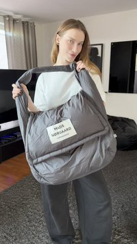Geantă de umăr căptușită gri, cu corp dreptunghiular, având o etichetă albă cu inscripția „MADS NØRGAARD COPENHAGEN.” Textură fină și moale.