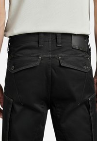 Bakomvy av person som bär svarta cargobyxor med knäppta ficklock och en vit inskjortad skjorta.