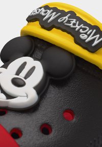 Μαύρο και κόκκινο ξύλινο παπούτσι με ανάγλυφο το πρόσωπο του Μίκυ Μάους και κίτρινο λουράκι που φέρει το κείμενο "Mickey Mouse" σε άσπρο χρώμα πάνω σε μαύρο φόντο.