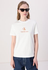 Valkoinen puuvillapaita, jossa on oranssi Calvin Klein Jeans -logo keskustassa. Mallilla on päällään tummat aurinkolasit ja löysät farkut.