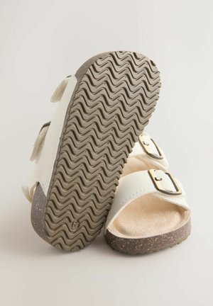 Valged sandaalid, millel on kaks reguleeritavat rihma metallist pannaldega, pruunimustrilise kummist tallaga ja pehme beež sisemusega.