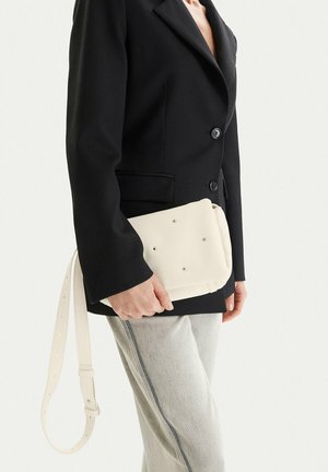 Person in schwarzem Blazer und hellen Jeans hält eine weiße rechteckige Handtasche mit Metallnieten und einem langen herunterhängenden Riemen.