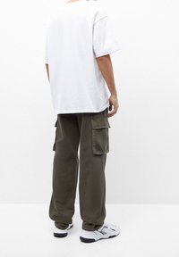 Person steht mit dem Rücken zur Kamera und trägt ein oversized weißes T-Shirt, olivgrüne Cargo-Hosen mit Seitentaschen und weiße Sneaker mit schwarzen Sohlen.