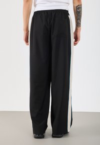 Melnas platās bikses ar teksturētu audumu, ar horizontālu krēmbaltu svītru un gaiši ziliem akcentiem sānos. Elastīgs jostasvieta, aizmugurējā kabata.