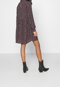Schwarzes Kleid mit floralem Muster, hohem Kragen, langen Ärmeln und Schlüsselloch-Rücken, kombiniert mit schwarzen Knöchelstiefeln mit klobigen Absätzen.