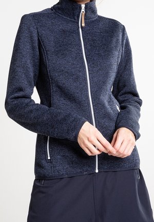 Femme portant une veste polaire zippée intégralement de couleur bleu foncé avec des poches zippées, debout les mains jointes à la taille.