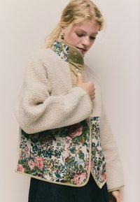 Beige fleecejakke med blomsterstof panel; har en kabelkrave og delikat piping, der giver en blød tekstur og et levende mønster.