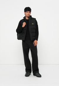 Black padded vest with high collar, black long-sleeve top, and loose black pants. Teamed with a black backpack and patterned black shoes.