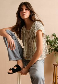 Blouse blanche à motifs floraux avec des manches courtes, design plissé. Associée à un pantalon court bleu clair et des sandales noires à glissière avec des accents dorés.