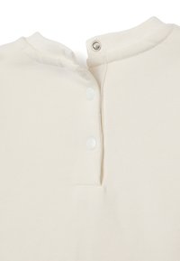 Cremfarbener, strukturierter Stoff mit Rundhalsausschnitt und drei weißen Druckknöpfen hinten zum Verschließen. Sauberes, minimalistisches Design.