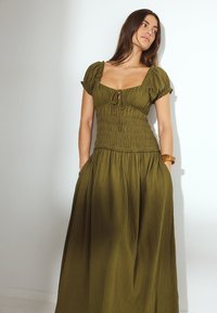 Olivenfarbiges Kleid mit Puffärmeln, gerafftem Oberteil und Bindeband am Ausschnitt. Volle, fließende Rock und seitliche Taschen. Strukturierter Stoff.