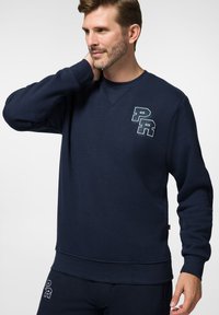 Granatowy sweter z teksturowanej tkaniny, z okrągłym dekoltem, długimi rękawami i haftowanym logo "PR" na piersi.