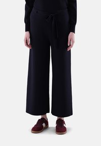 Weit geschnittene, dunkelblaue Hosen aus weichem Material mit einem vorderen Gürtel und tailliertem Bund. Kombiniert mit burgunderfarbenen Sneakers mit weißen Akzenten.