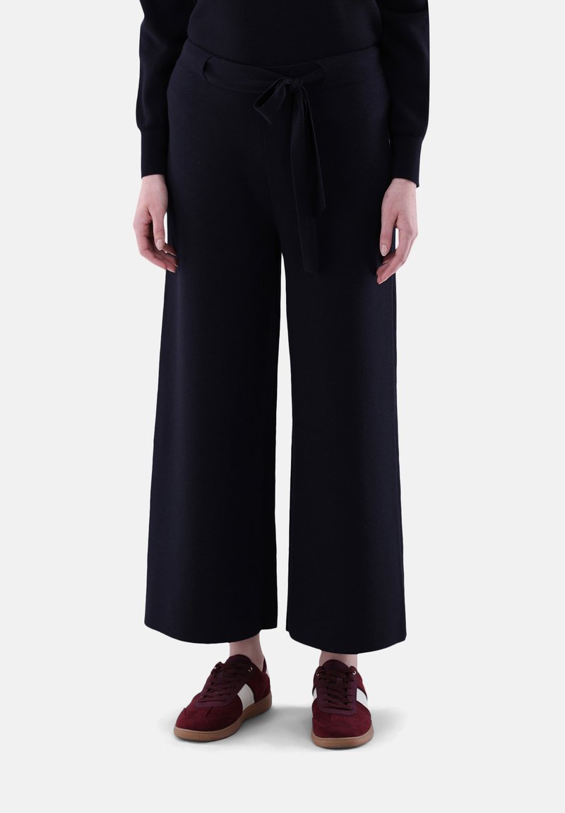 Weit geschnittene, dunkelblaue Hosen aus weichem Material mit einem vorderen Gürtel und tailliertem Bund. Kombiniert mit burgunderfarbenen Sneakers mit weißen Akzenten.