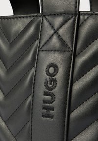 Detalle de bolsa de cuero negro con costuras en forma de chevron y nombre de la marca "HUNGO" bordado en negro en la correa vertical.