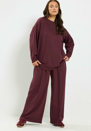 Donna in piedi che indossa una felpa bordeaux larga, pantaloni ampi abbinati e sneakers bordeaux su uno sfondo chiaro e semplice.