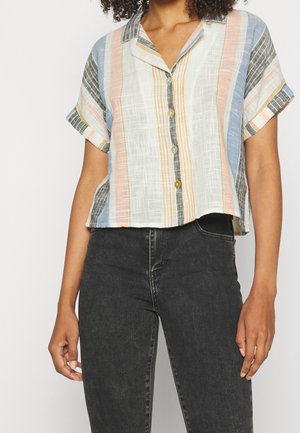 Kvinde iført en kortærmet, knappet skjorte med lodrette pastelfarvede striber, kombineret med højtaljede sorte jeans.