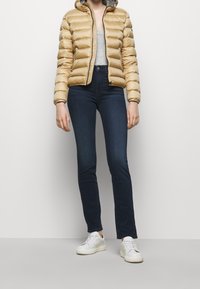 Gouden donsjas met capuchon, ritssluiting aan de voorkant en doorgestikte structuur. Gecombineerd met donkere skinny jeans en witte sneakers.