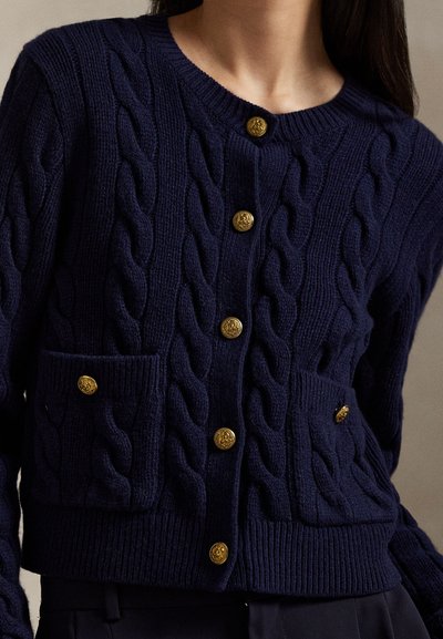 Navyblå kabelstrikket cardigan med guldknapper og to forlommer på en person med langt mørkt hår.